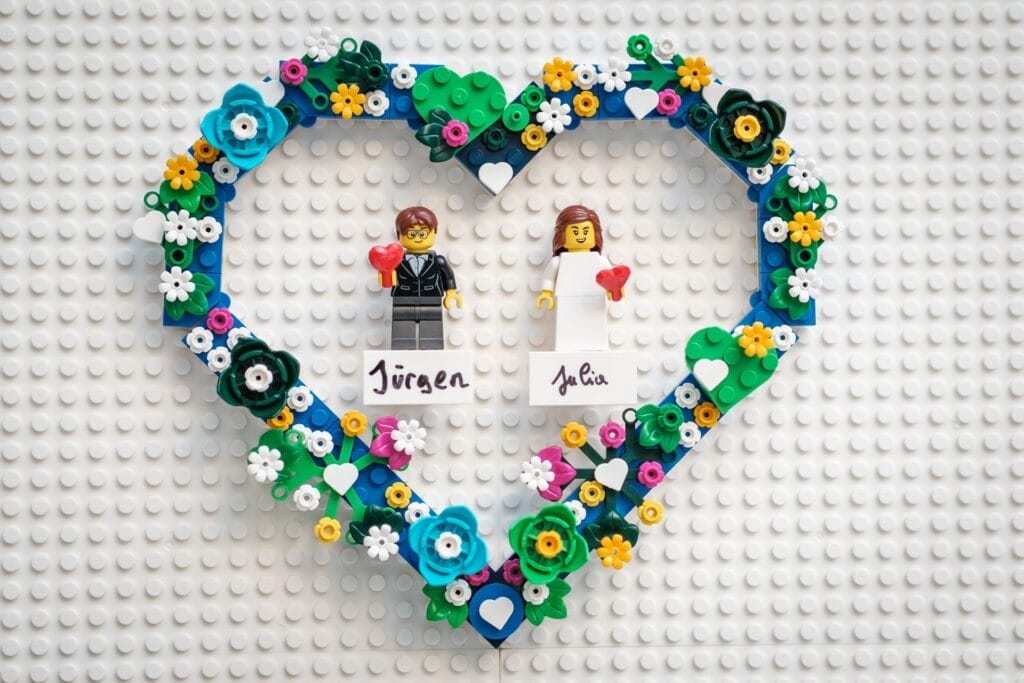 Hochzeitskrone Dieser bezaubernde Lego-Herzrahmen fängt bezaubernde Momente mit zwei Figuren ein, die rote Herzen halten, umgeben von bunten Lego-Blumen. Darunter feiern die Namen „Jürgen“ und „Julia“ ihre Verbundenheit in einer entzückenden Miniaturwelt.