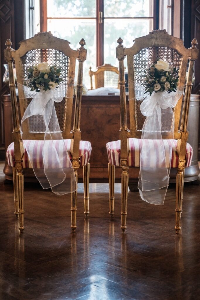 Hochzeitskrone Zwei verzierte Holzstühle mit geflochtenen Rückenlehnen, geschmückt mit weißen Blumen und transparenten Bändern, stehen auf einem polierten Boden vor einem Fenster und sind bereit, bezaubernde Momente in natürlichen Hochzeitsfotos festzuhalten.