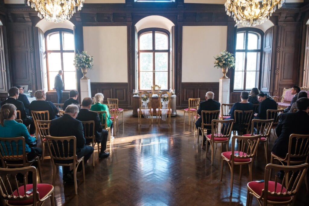 Hochzeitskrone In einem formellen Raum mit großen Fenstern und Kronleuchtern sitzen Menschen auf Stühlen, mit Blick auf einen zentralen Bereich, der mit Blumenarrangements und zwei leeren, reich verzierten Stühlen geschmückt ist, und sind bereit, bezaubernde Momente festzuhalten, die an Hochzeitsfotografie in Niederösterreich erinnern.