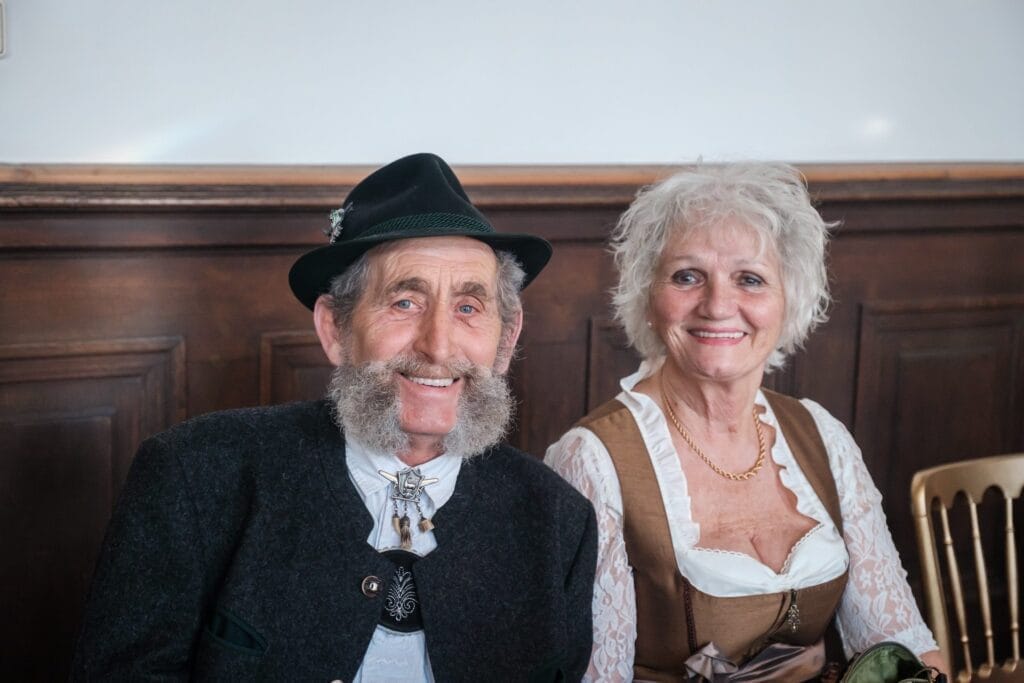 Hochzeitskrone Ein älterer Mann und eine ältere Frau, beide in traditioneller bayerischer Kleidung gekleidet, werden in bezaubernden Momenten festgehalten, als sie zusammensitzen und in die Kamera lächeln.