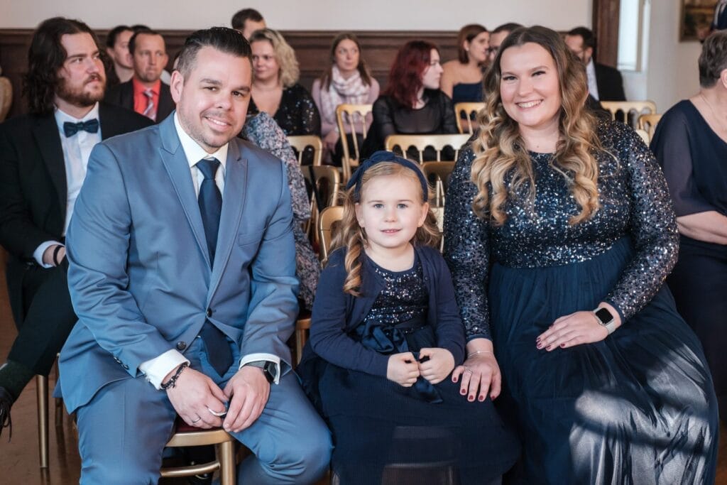 Hochzeitskrone In einem formellen Rahmen sitzen ein Mann und eine Frau in passenden blauen Anzügen neben einem jungen Mädchen in Blau. Andere Gäste sitzen im Hintergrund und fangen einen jener bezaubernden Momente ein, die man oft in der Hochzeitsfotografie in Niederösterreich findet.