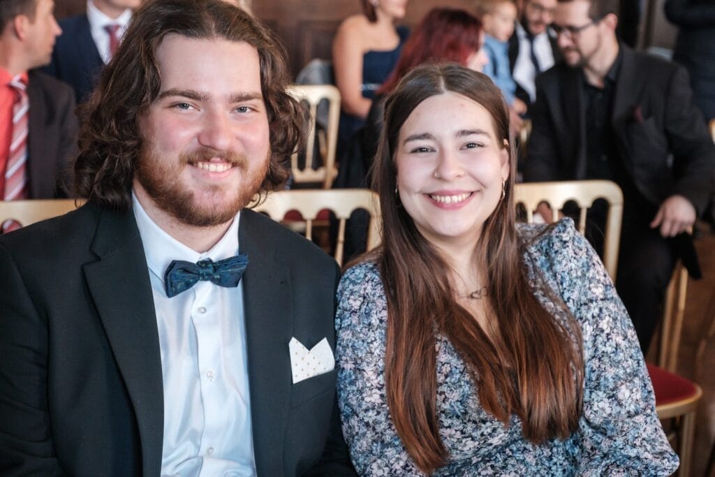 Hochzeitskrone Zwei Personen sitzen und lächeln bei einer formellen Veranstaltung, umgeben von anderen Gästen. Der Mann trägt einen Anzug mit Fliege und die Frau ein geblümtes Kleid und fängt bezaubernde Momente ein. Perfekt für alle, die natürliche Hochzeitsfotos suchen, die eine Geschichte von Freude und Eleganz erzählen.