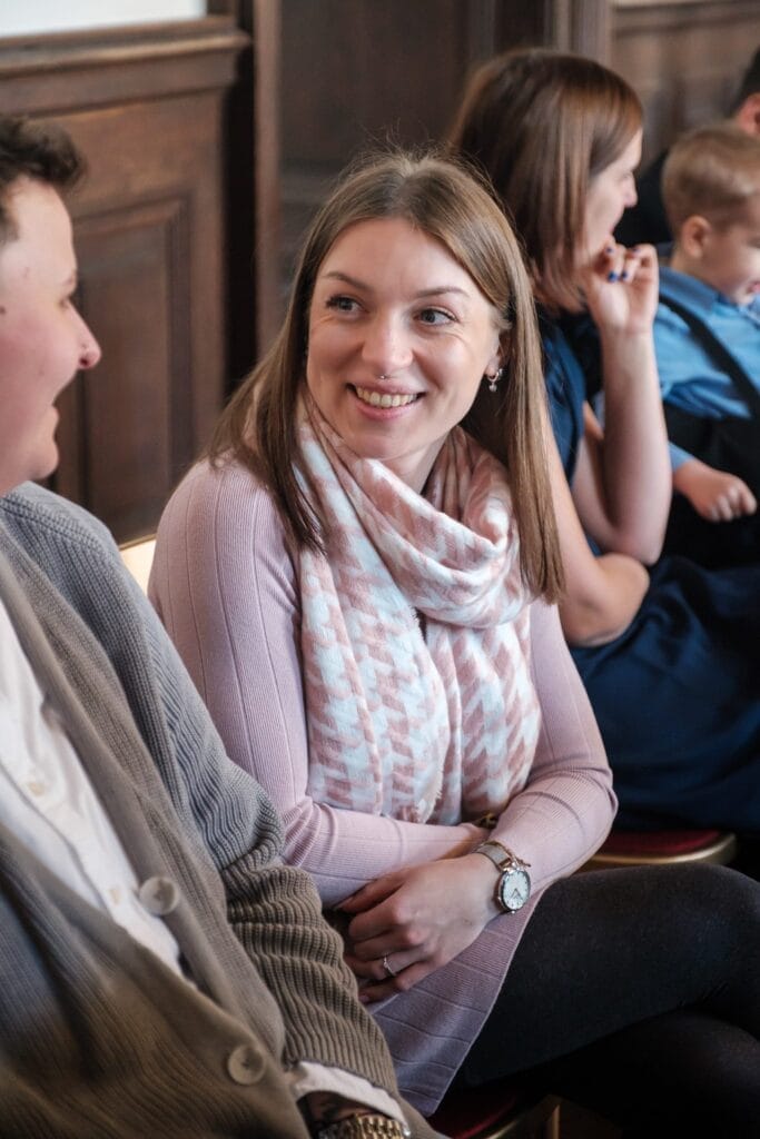 Hochzeitskrone Eine Frau in einem rosa Pullover und Schal sitzt lächelnd da und unterhält sich mit einer Person zu ihrer Linken. Hinter ihr scheinen mehrere Personen, darunter ein Kind, die bezaubernden Momente zu genießen, die wie auf natürlichen Hochzeitsfotos festgehalten wurden.