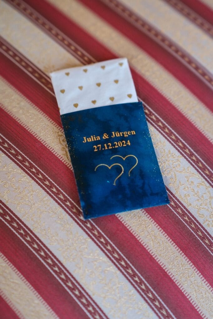 Hochzeitskrone Ein Hochzeitsgeschenk ziert eine gemusterte rot-goldene Tischdecke, mit der Aufschrift „Julia & Jürgen, 27.12.2024“ und goldenen Herzen auf einer blauen Tasche, und fängt bezaubernde Momente ein, die in der Hochzeitsfotografie in Niederösterreich glänzen würden.