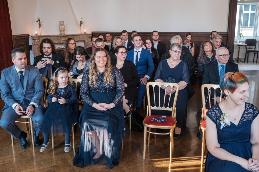 Hochzeitskrone Eine Gruppe von Menschen, die in einem Innenbereich sitzen und für eine Veranstaltung formell gekleidet sind, wurde in bezaubernden Momenten festgehalten. Eine Mischung aus Erwachsenen und einem Kind blickt in die Kamera, die Stühle sind in einem strukturierten Layout angeordnet, das an Hochzeitsfotografie in Niederösterreich erinnert.