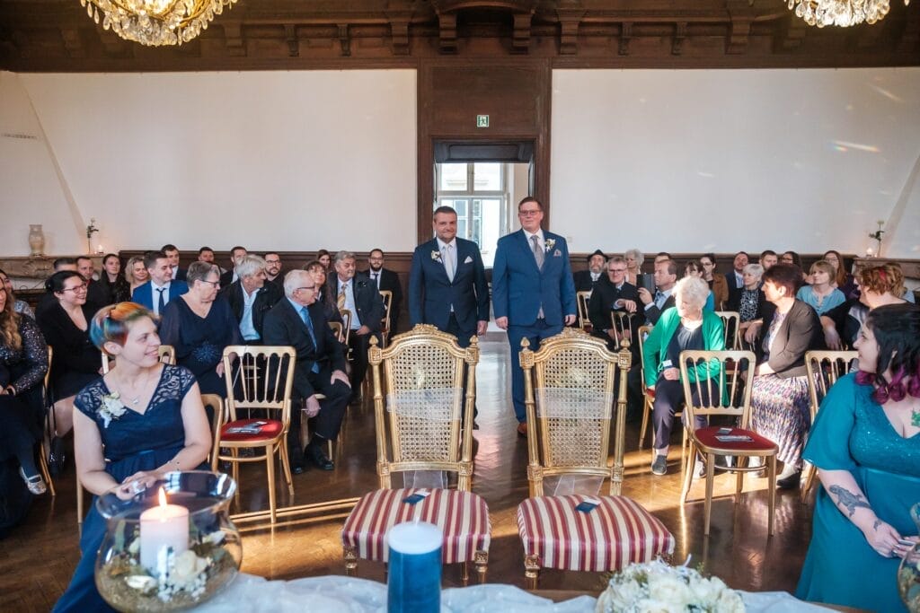 Hochzeitskrone Zwei Bräutigame stehen vor ihren Gästen in einem eleganten Raum mit Kronleuchtern und reich verzierten Stühlen und bereiten sich auf eine Zeremonie vor, die von natürlichen Hochzeitsfotos festgehalten wurde.