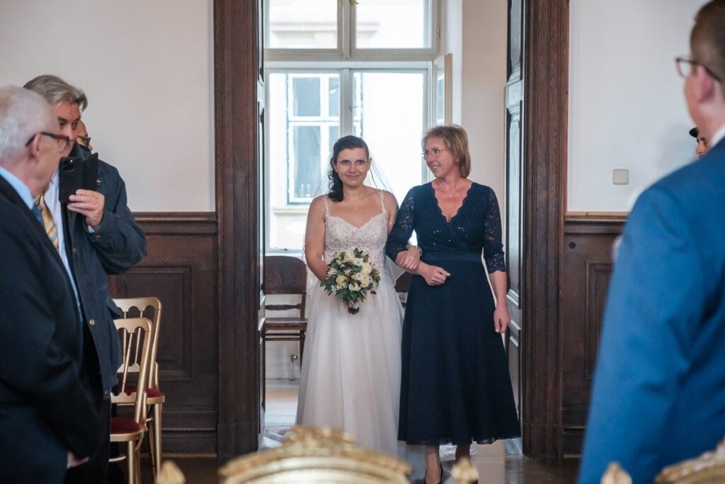 Hochzeitskrone Eine Braut betritt mit einem Blumenstrauß in der Hand einen Raum, begleitet von einer Frau. Mehrere Personen sitzen und stehen und beobachten in dem holzgetäfelten Raum mit einem Fenster im Hintergrund – bezaubernde Momente der Hochzeitsfotografie in Niederösterreich.