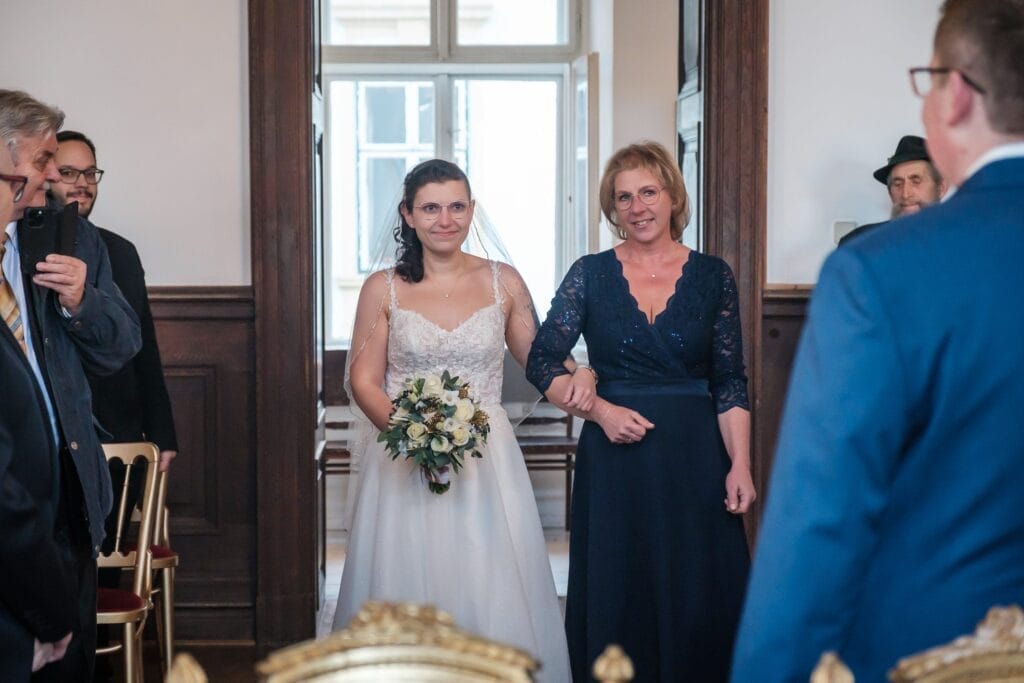 Hochzeitskrone Eine Braut mit Brille schreitet in einem weißen Kleid den Gang entlang und hält einen Blumenstrauß in der Hand. Sie wird von einer Frau in Blau begleitet, die bezaubernde Momente festhält. Die Gäste schauen in einem Raum mit Holzvertäfelung zu, der perfekt für natürliche Hochzeitsfotos ist, die die Schönheit der Hochzeitsfotografie in Niederösterreich hervorheben.