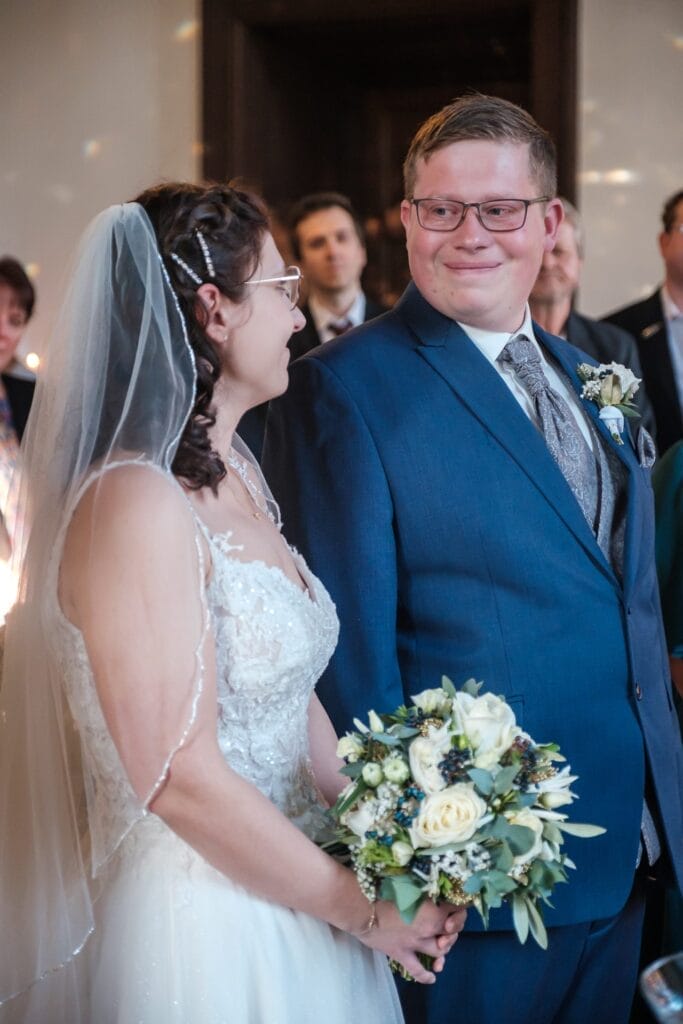 Hochzeitskrone Eine Braut in einem weißen Kleid und Schleier hält einen Strauß weißer Blumen und lächelt ihren Bräutigam im blauen Anzug während der Hochzeitszeremonie an und fängt bezaubernde Momente natürlicher Hochzeitsfotos ein.