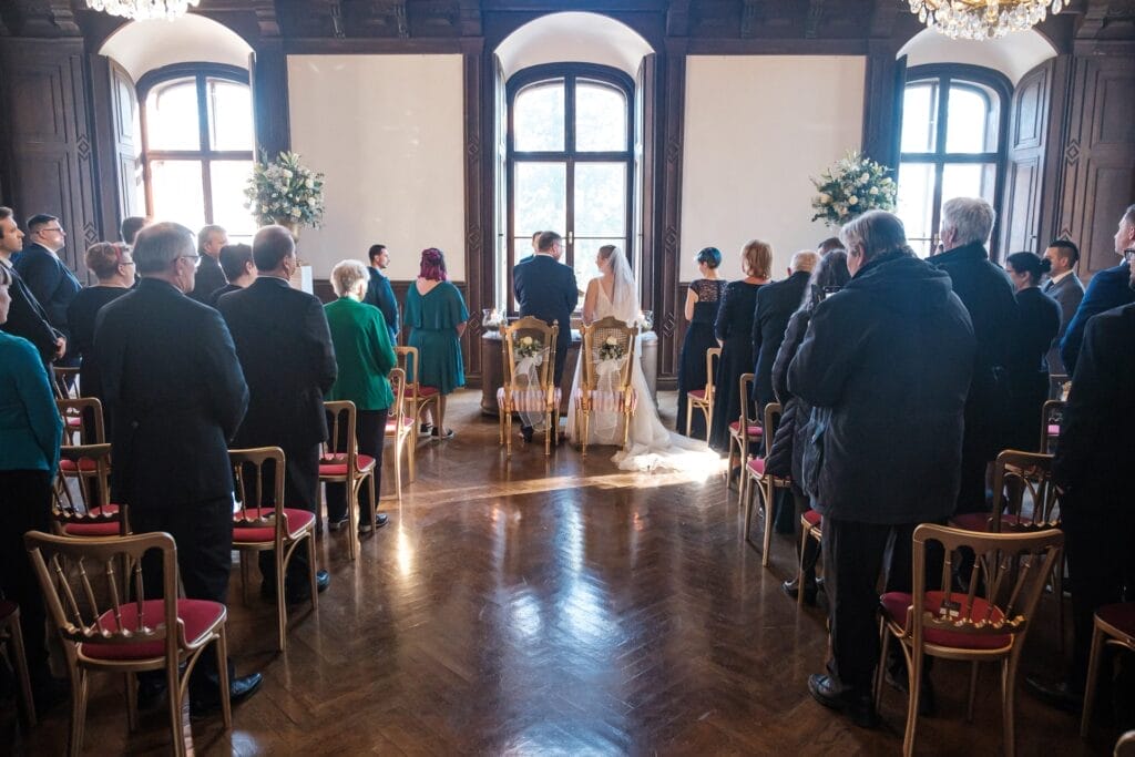 Hochzeitskrone Eine Hochzeitszeremonie, die in natürliches Licht getaucht ist, das durch große Fenster fällt. Das Paar sitzt dem Geistlichen gegenüber, umgeben von stehenden Gästen. Blumenarrangements flankieren die Fenster und fangen bezaubernde Momente ein. Stühle mit roten Kissen verleihen dieser bezaubernden Szene eine lebendige Note.