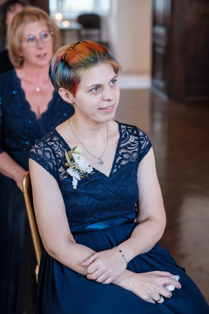 Hochzeitskrone Eine Person mit bunten Kurzhaarhaaren und Blumenanstecker sitzt in einem blauen Spitzenkleid und hält bezaubernde Momente bei einem Indoor-Event fest.