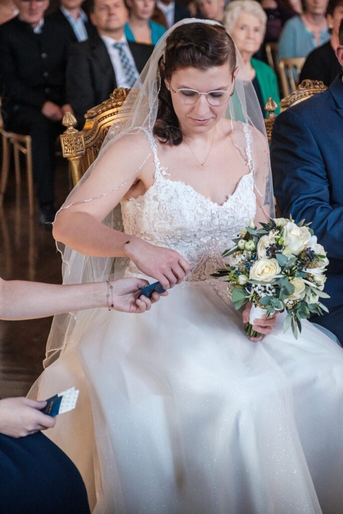 Hochzeitskrone Eine Braut in weißem Kleid und Schleier hält einen Blumenstrauß und hält mit einem Mikrofon bezaubernde Momente fest. Im Hintergrund sitzen Gäste und unterstreichen den natürlichen Charme der Hochzeitsfotografie in Niederösterreich.