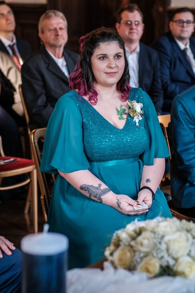 Hochzeitskrone Eine Frau in einem blaugrünen Kleid sitzt bei einer Veranstaltung und hält eine Notiz in der Hand. Geschmückt mit einer Blumenbrosche und sichtbaren Tattoos ist sie von anderen Personen in formeller Kleidung umgeben und fängt bezaubernde Momente ein, die an natürliche Hochzeitsfotos erinnern.