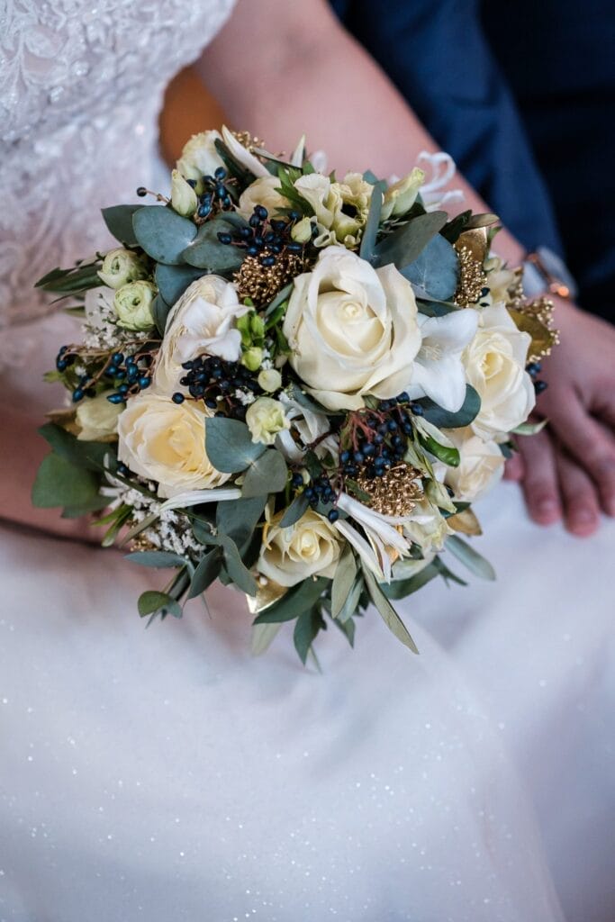 Hochzeitskrone Nahaufnahme einer Braut, die einen Blumenstrauß mit weißen Rosen, Grünpflanzen und kleinen dekorativen Akzenten hält und bezaubernde Momente einfängt, während sie neben einer Person in einem dunklen Anzug sitzt.