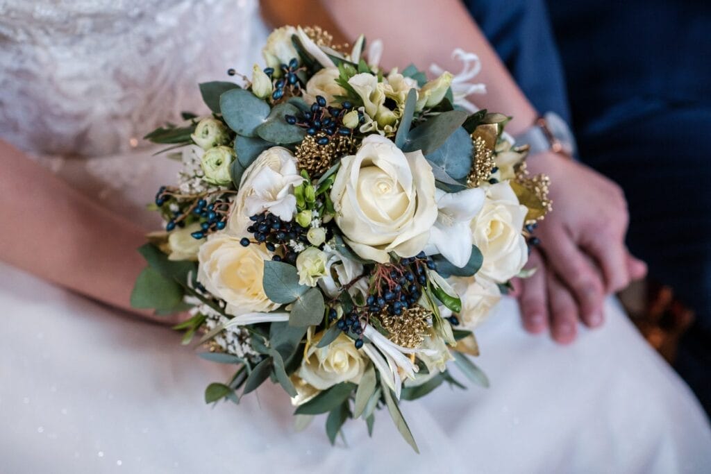 Hochzeitskrone Nahaufnahme eines Brautstraußes mit weißen Rosen, Grünpflanzen und kleinen dunklen Beeren, gehalten von einer Person in einem weißen Spitzenkleid, die neben jemandem in einem dunklen Anzug sitzt. Zauberhafte Momente, perfekt festgehalten in natürlichen Hochzeitsfotos, sind unvergänglich.