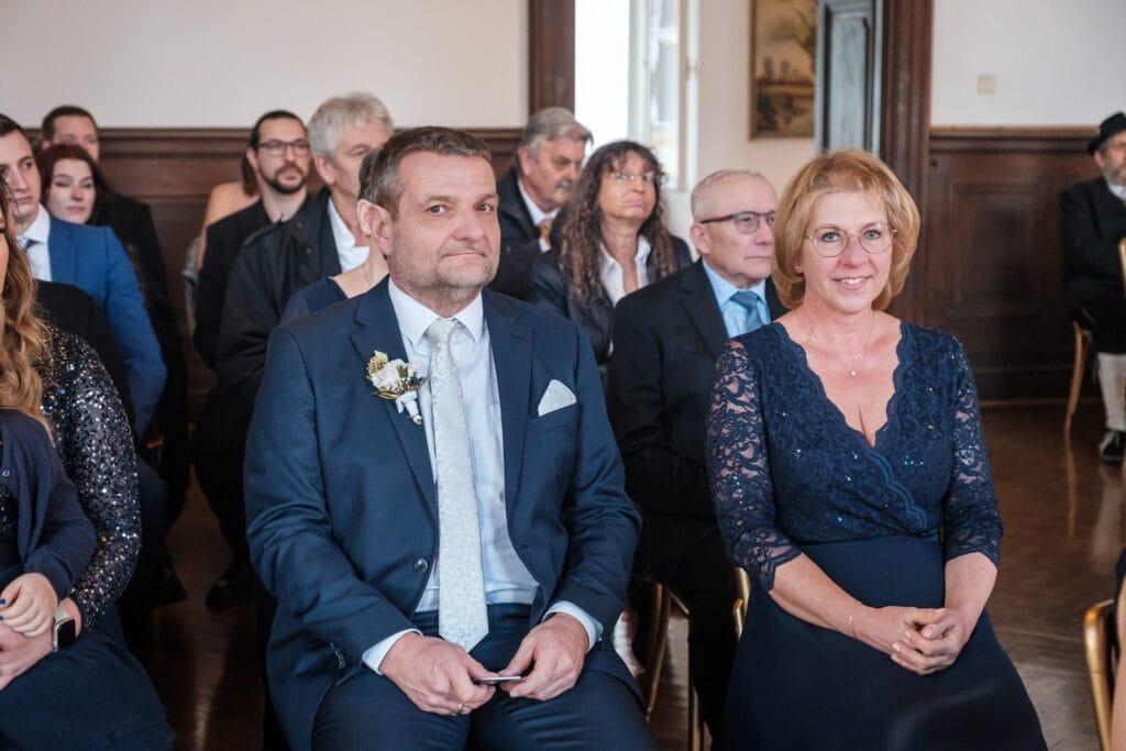 Hochzeitskrone In einem formellen Rahmen versammelt sich eine Gruppe von Menschen, wobei ein Mann und eine Frau im Vordergrund, elegant in Blau gekleidet, bezaubernde Momente teilen. Mit einem herzlichen Lächeln verkörpern sie den Charme natürlicher Hochzeitsfotos, der typisch für exquisite Hochzeitsfotografie in Niederösterreich ist.