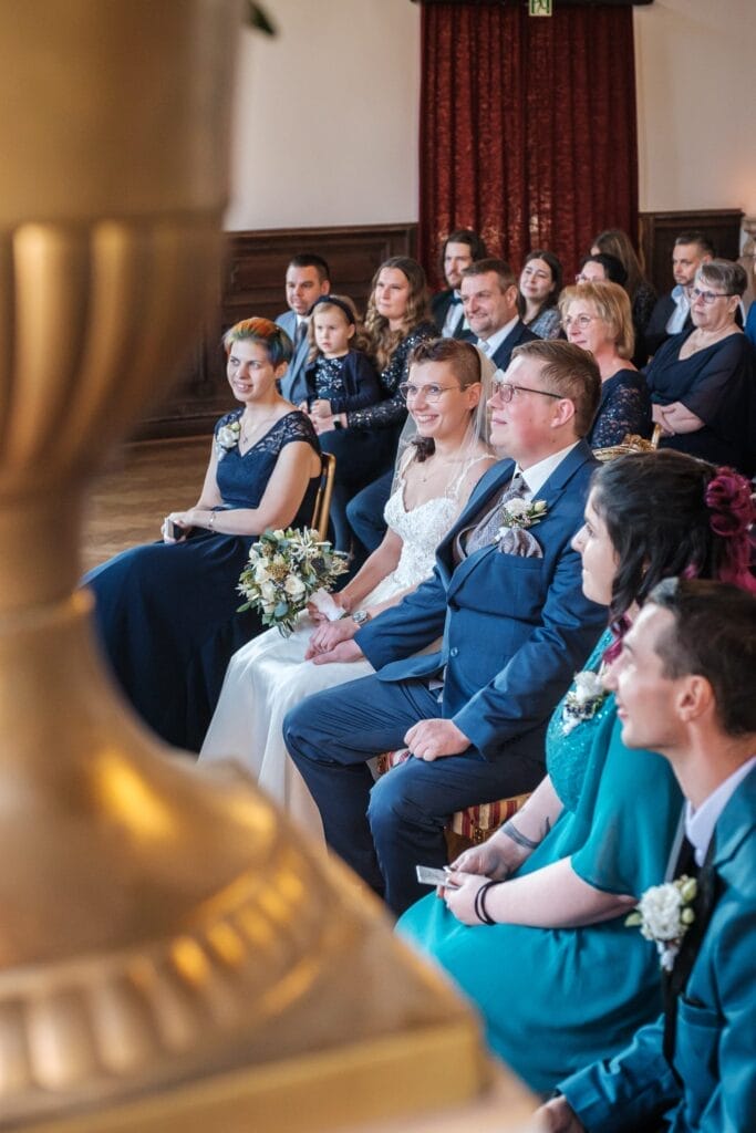 Hochzeitskrone Während der Zeremonie sitzen Braut und Bräutigam zusammen, umgeben von Gästen, die von dem Moment fasziniert sind. Die Braut hält einen Blumenstrauß, der so bezaubernd ist wie jeder andere in der Hochzeitsfotografie in Niederösterreich, und alle konzentrieren sich nach vorne.