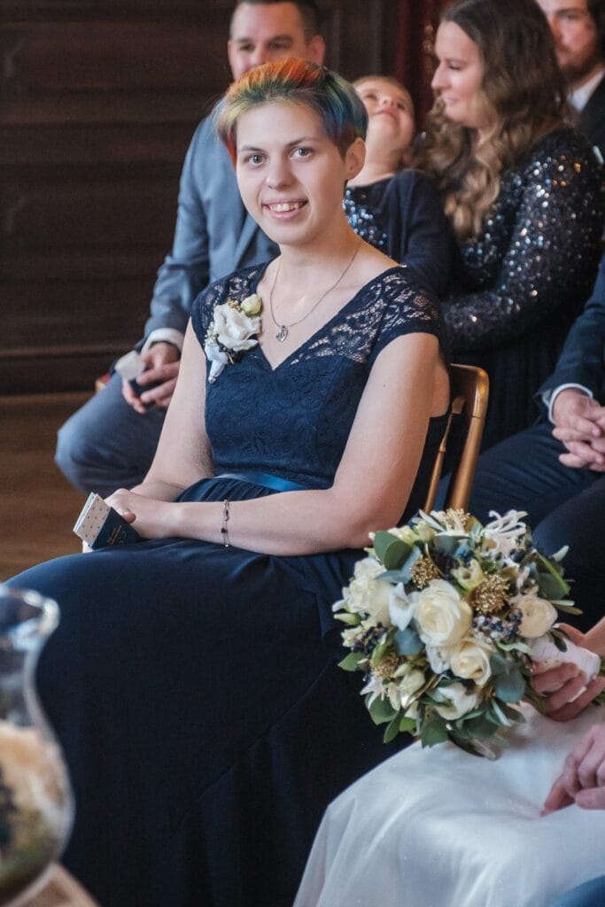 Hochzeitskrone Eine Person in einem dunkelblauen Spitzenkleid mit Blumenanstecker sitzt auf einem Stuhl, hält Papiere in der Hand und fängt bezaubernde Momente ein. Ein Blumenstrauß und zwei Personen in dunkler Kleidung vervollständigen diese Szene natürlicher Hochzeitsfotos, wunderschön gerahmt durch die Kunst der Hochzeitsfotografie in Niederösterreich.