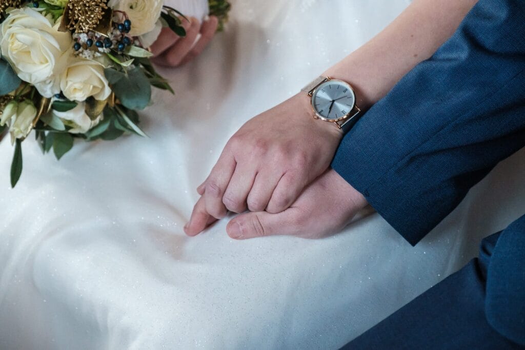 Hochzeitskrone Zwei ineinander verschlungene Hände, einer trägt eine Uhr. Ein Strauß weißer Rosen und Grünpflanzen ruht daneben auf einem weißen Stoff und fängt bezaubernde Momente in diesem natürlichen Hochzeitsfotos ein.