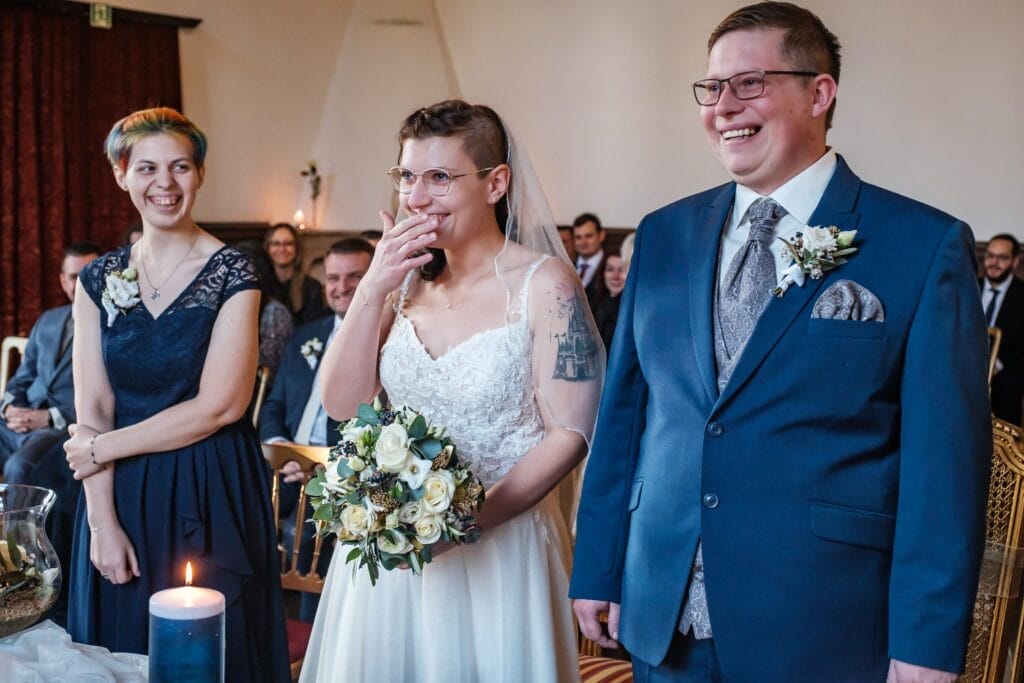 Hochzeitskrone Braut und Bräutigam stehen lächelnd in einem Raum, wobei die Braut einen Blumenstrauß hält. Neben ihnen trägt eine Brautjungfer in einem blauen Kleid zu den bezaubernden Momenten bei. Im Hintergrund sitzen die Gäste und machen natürliche Hochzeitsfotos, die ihren besonderen Tag hervorheben.