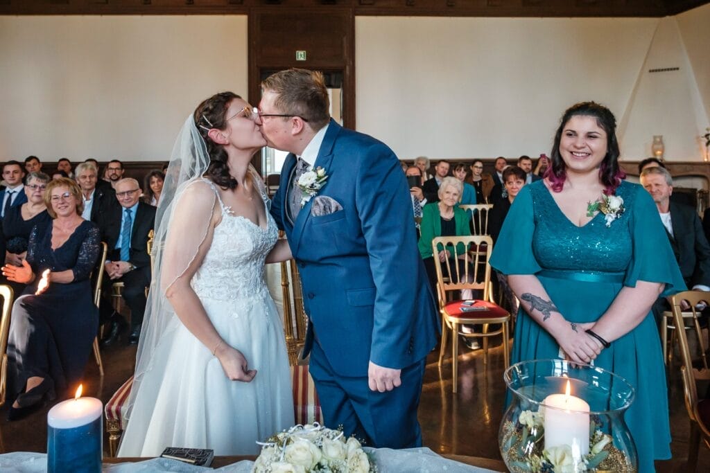 Hochzeitskrone Braut und Bräutigam küssen sich bei ihrer Hochzeitszeremonie und halten bezaubernde Momente fest. Im Hintergrund sitzen die Gäste, daneben steht eine lächelnde Frau – ein perfektes Beispiel für natürliche Hochzeitsfotos in Niederösterreich.