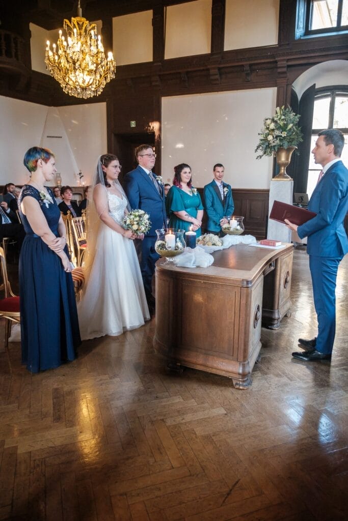 Hochzeitskrone In einem wunderschön dekorierten Raum findet eine Hochzeitszeremonie statt, während das Paar vor einem Geistlichen an einem Holztisch steht. Die Trauzeugen versammeln sich in der Nähe, während die Gäste zuschauen und diese bezaubernden Momente genießen. Perfekt für natürliche Hochzeitsfotos in Niederösterreich.