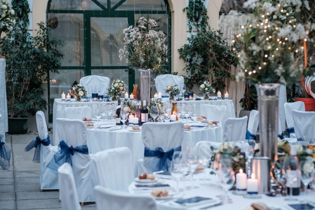 Hochzeitskrone Ein eleganter Empfangsbereich mit runden Tischen, weißen Tischdecken und dekorativen Tafelaufsätzen. Jeder Tisch ist mit Kerzen, Glaswaren und Blumenarrangements gedeckt, umgeben von Grün – perfekt, um bezaubernde Momente in der Hochzeitsfotografie in Niederösterreich festzuhalten.