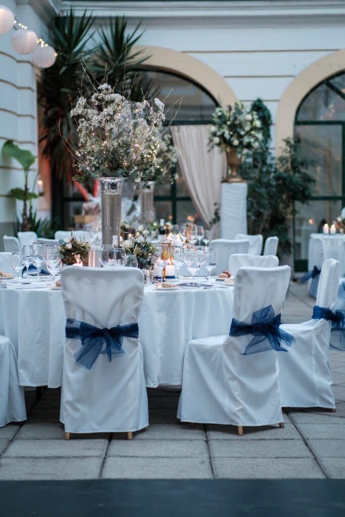 Hochzeitskrone Erleben Sie bezaubernde Momente bei einer eleganten Hochzeitsfeier im Freien, bei der runde Tische mit weißen Tischdecken und blauen Stuhlschleifen geschmückt sind. Hohe Glasvasen mit zarten Blumen dienen als Tischdekoration, umgeben von Kerzen und Grünpflanzen – bezaubernd für jede Hochzeitsfotografie in Niederösterreich.