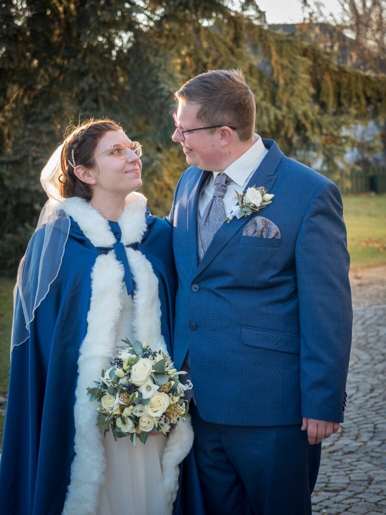 Hochzeitskrone Ein Paar, in formelle Kleidung mit Blumen gekleidet, steht auf einem Steinweg und lächelt sich an. Die Frau trägt einen Schleier und einen Umhang, während der Mann einen blauen Anzug trägt. Im Hintergrund sind Bäume zu sehen, die bezaubernde Momente natürlicher Hochzeitsfotos einfangen.