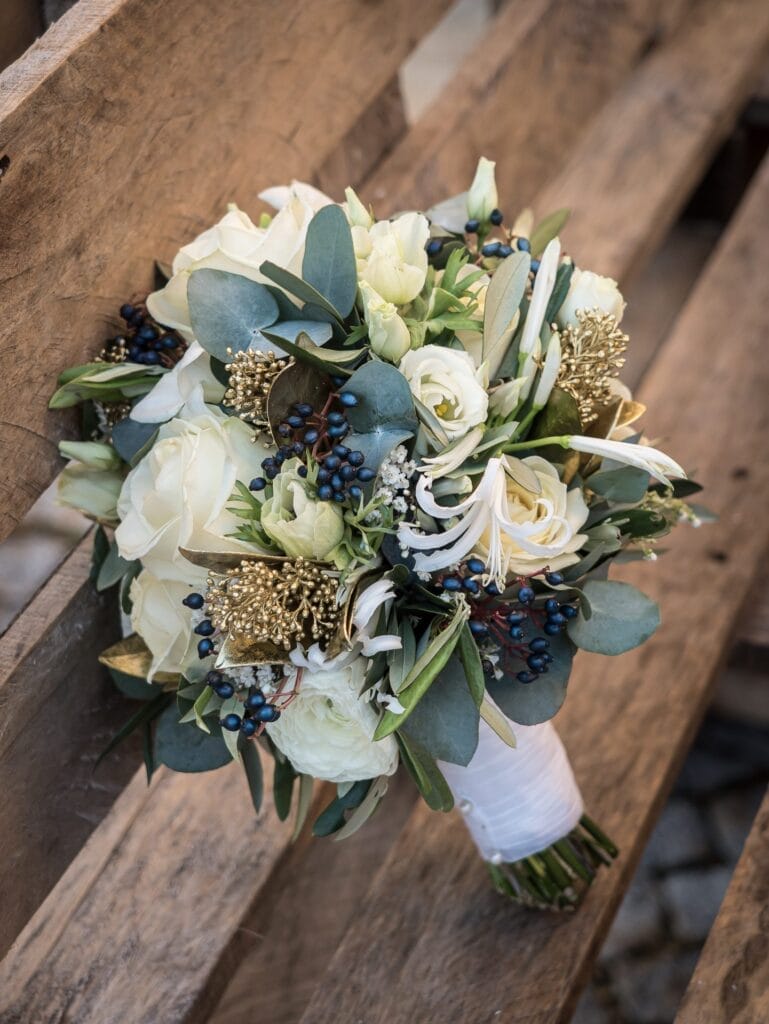 Hochzeitskrone Ein Strauß weißer Rosen, Grünpflanzen und Trockenblumen, zusammengebunden mit einer weißen Schleife, ruht auf einer Holzbank – eine perfekte Note für bezaubernde Momente, festgehalten in der Hochzeitsfotografie in Niederösterreich.