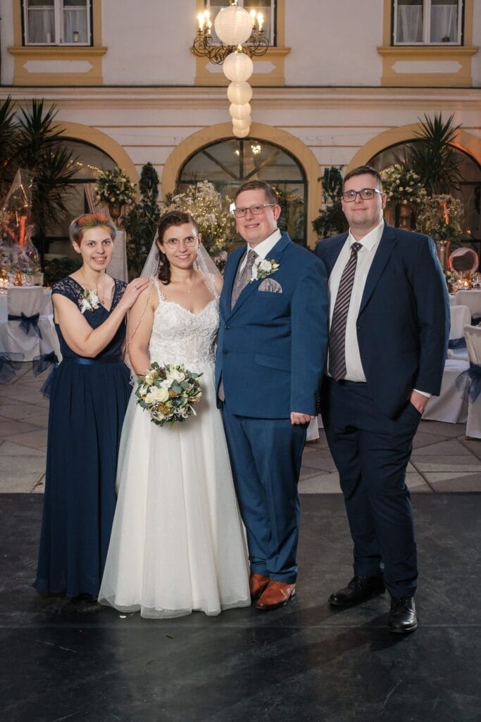 Hochzeitskrone Vier Personen posieren zusammen in Innenräumen für Hochzeitsfotografie in Niederösterreich; eine Braut in einem weißen Kleid mit Blumen in der Hand, ein Bräutigam in einem blauen Anzug und zwei weitere in Abendgarderobe, alle lächeln und schaffen bezaubernde Momente.