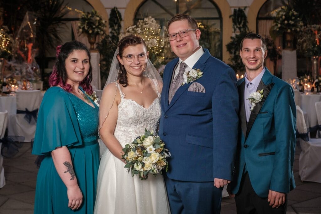 Hochzeitskrone Vier Personen in festlicher Kleidung posieren gemeinsam bei einer Hochzeit und fangen bezaubernde Momente ein. Braut und Bräutigam stehen in der Mitte, flankiert von zwei Trauzeugen. Der Hintergrund ist mit Blumen und Tischen geschmückt und schafft eine perfekte Szene für Hochzeitsfotografie in Niederösterreich.