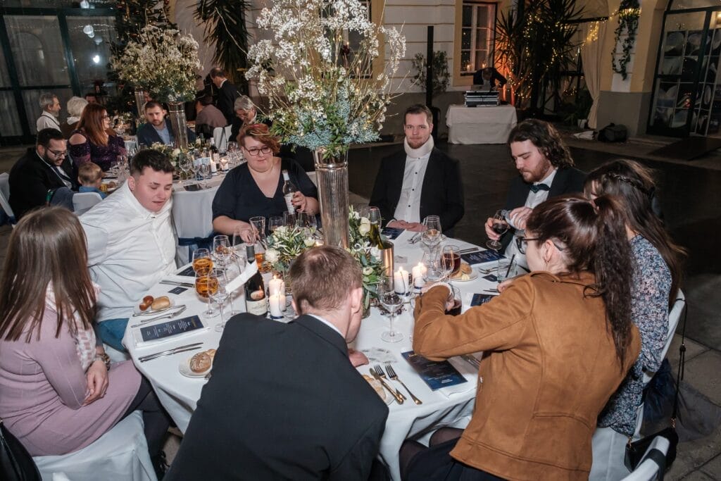 Hochzeitskrone Menschen sitzen bei einer Veranstaltung in geschlossenen Räumen um einen formell gedeckten Tisch mit Blumenschmuck und brennenden Kerzen und fangen bezaubernde Momente ein, die sich perfekt für natürliche Hochzeitsfotos eignen.