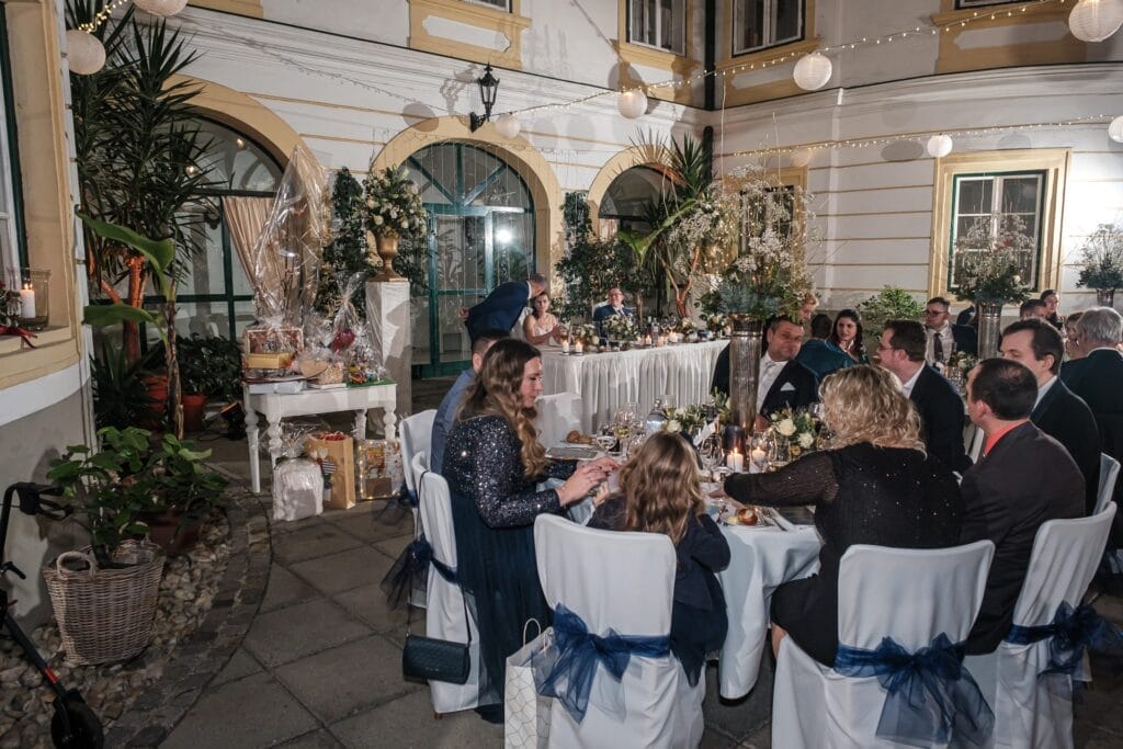 Hochzeitskrone Gäste sitzen an Tischen mit weißen Tischdecken und Stühlen, genießen ein Essen bei einer Abendveranstaltung in einem wunderschön dekorierten Innenhof und fangen bezaubernde Momente ein, die den Charme natürlicher Hochzeitsfotos widerspiegeln.