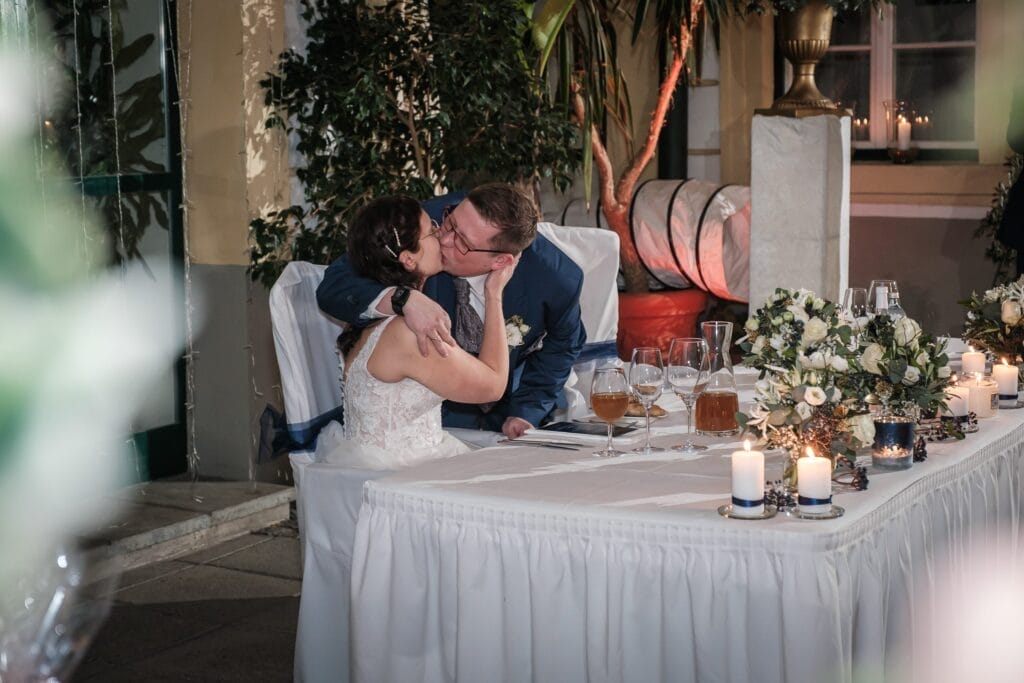 Hochzeitskrone An einem wunderschön dekorierten Hochzeitsempfangstisch, geschmückt mit Blumen und Kerzen, küsst sich ein Paar zärtlich und hält bezaubernde Momente ihres besonderen Tages fest. Mit natürlichen Hochzeitsfotos werden diese herzlichen Erinnerungen aus Niederösterreich für immer bewahrt.