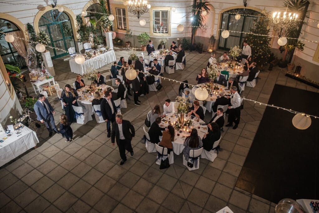 Hochzeitskrone Luftaufnahme einer formellen Innenveranstaltung mit Gästen an runden Tischen, die bezaubernde Momente einfangen. Hängelampen und Kronleuchter verleihen dem Raum Eleganz, perfekt für Hochzeitsfotografie in Niederösterreich.