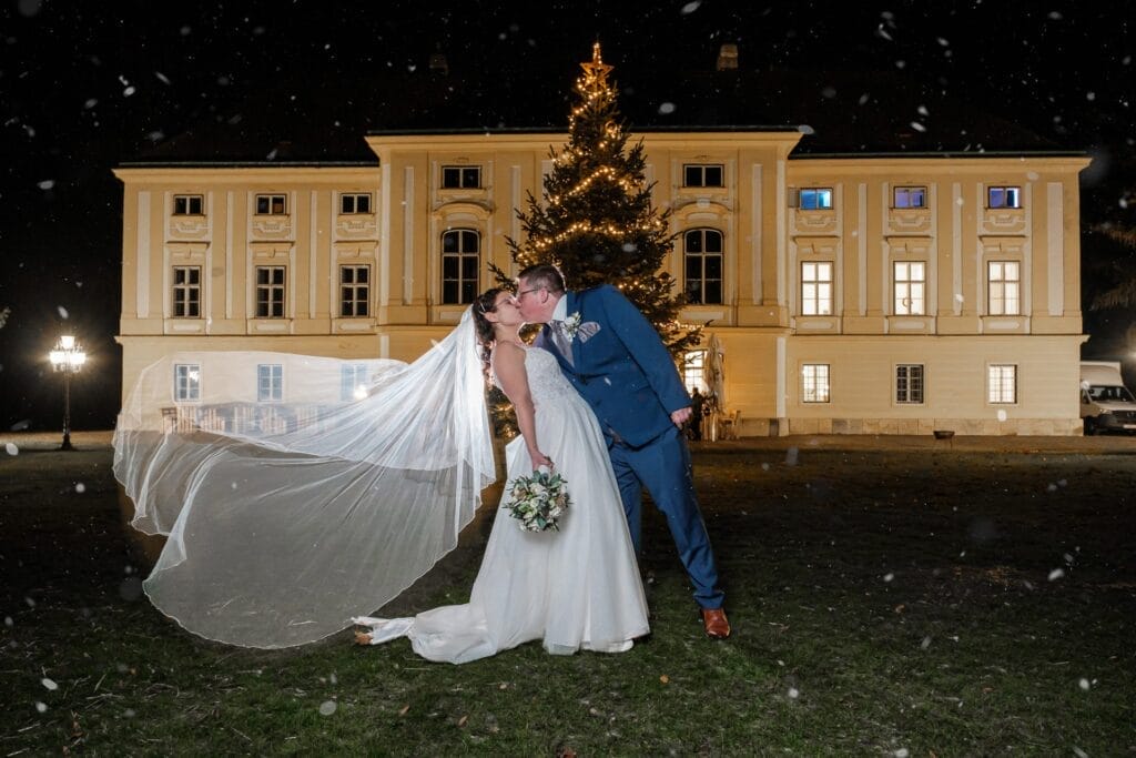 Hochzeitskrone Braut und Bräutigam küssen sich vor einem beleuchteten Gebäude in der Nacht, hinter ihnen verbreitet ein geschmückter Weihnachtsbaum ein festliches Licht. Der Schleier der Braut weht im Wind und fängt bezaubernde Momente ein, die Hochzeitsfotografie in Niederösterreich ausmachen.