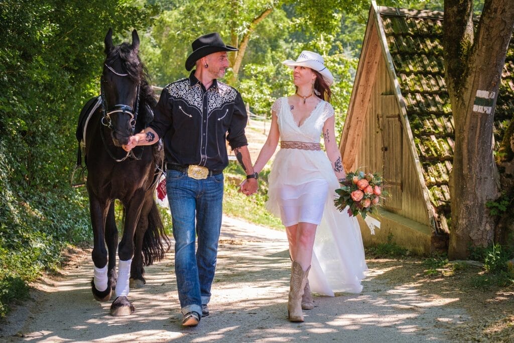 Hochzeitskrone Ein Paar in Westernkleidung spaziert Hand in Hand auf einem sonnigen Weg und führt ein schwarzes Pferd. Die Frau hält einen Blumenstrauß in der Hand und fängt damit echte Momente ein. Ihre Reise wird von einem Hochzeitsfotografen in Wien und Niederösterreich wunderschön dokumentiert.