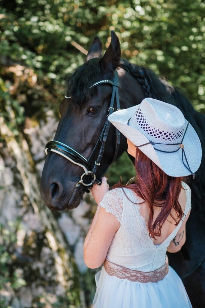 Hochzeitskrone Eine Frau in einem weißen Kleid und Cowboyhut streichelt ein schwarzes Pferd inmitten üppigen Grüns und fängt die Essenz natürlicher Hochzeitsfotos ein.