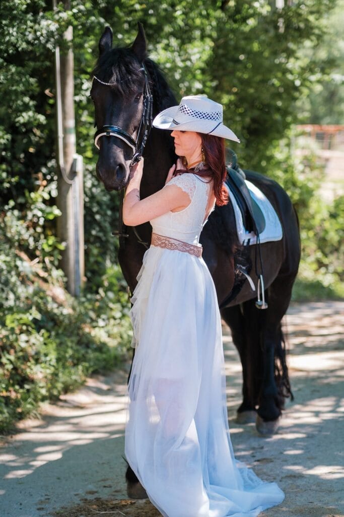 Hochzeitskrone Eine Frau in einem weißen Kleid und Hut posiert neben einem schwarzen Pferd im Freien in einem Waldstück und fängt die Essenz natürlicher Hochzeitsfotos ein.