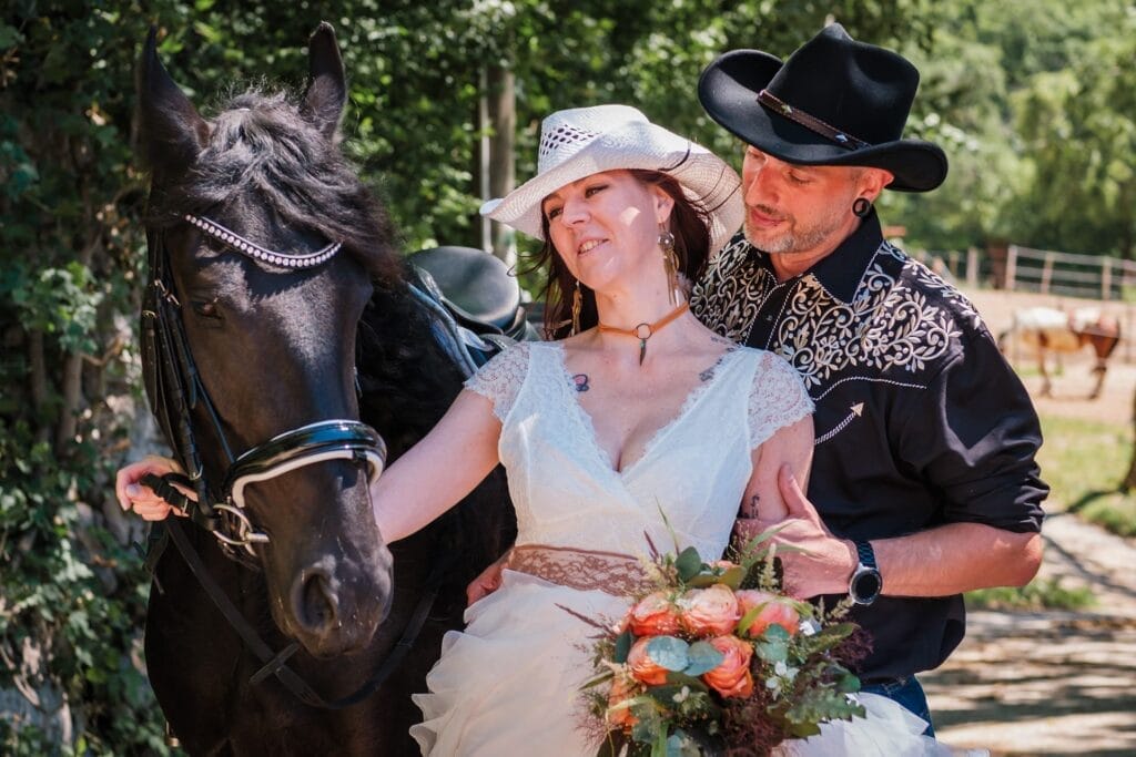 Hochzeitskrone Ein Paar in Westernkleidung posiert mit einem schwarzen Pferd und fängt echte Momente ein. Die Frau in weißem Kleid und Hut hält einen Blumenstrauß, während der Mann einen schwarzen Cowboyhut und ein besticktes Hemd trägt.