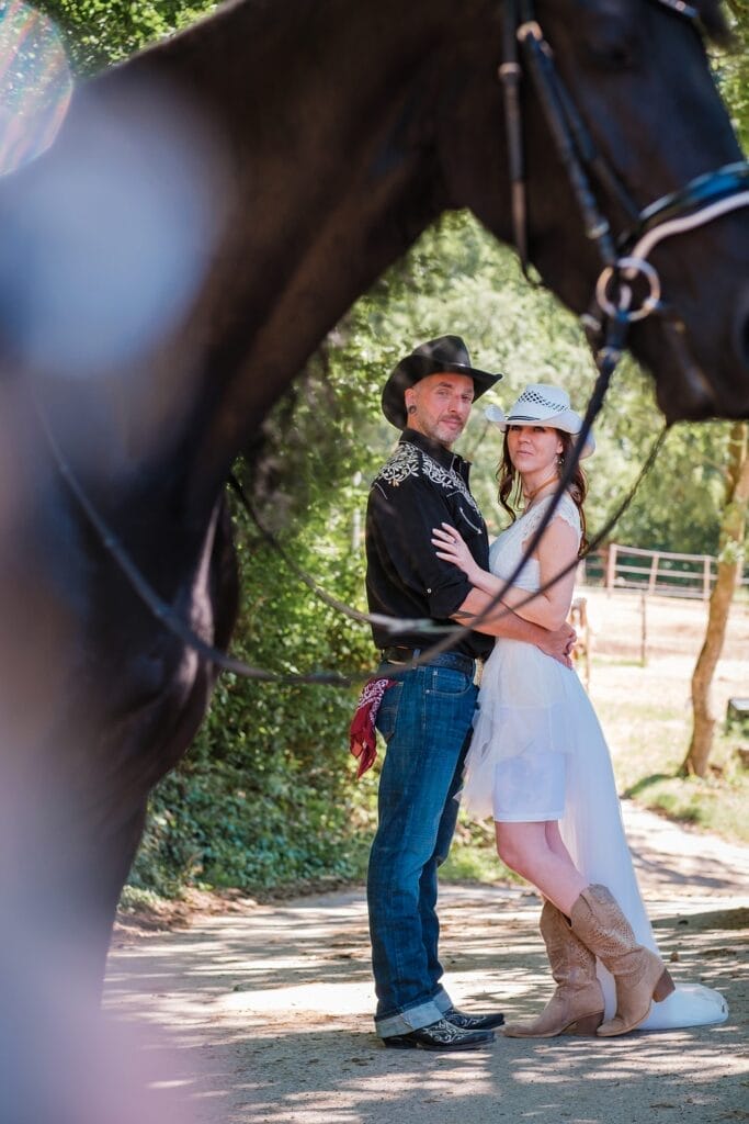 Hochzeitskrone Ein Mann und eine Frau in Cowboyhüten und -stiefeln stehen zusammen im Freien, eingerahmt vom Kopf und Hals eines schwarzen Pferdes, und fangen „echte Momente“ vor einer sonnigen, bewaldeten Kulisse ein.