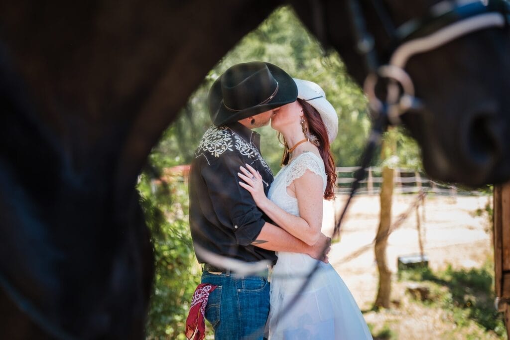 Hochzeitskrone Ein Paar mit Cowboyhüten küsst sich, umrahmt von zwei Pferden, im Freien an einem sonnigen Tag – echte Momente eingefangen.