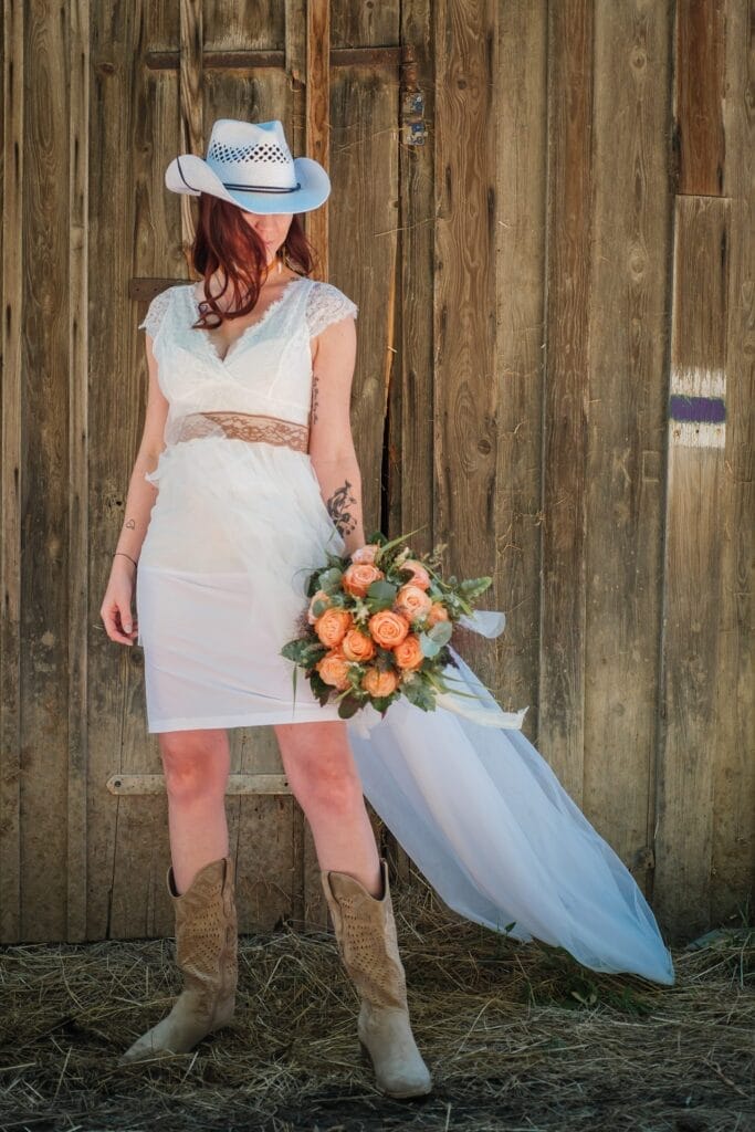 Hochzeitskrone Eine Person in einem weißen Kleid, Cowboyhut und Stiefeln steht mit einem Strauß orangefarbener Rosen vor einem hölzernen Hintergrund und fängt echte Momente für authentische Hochzeitsfotos ein.