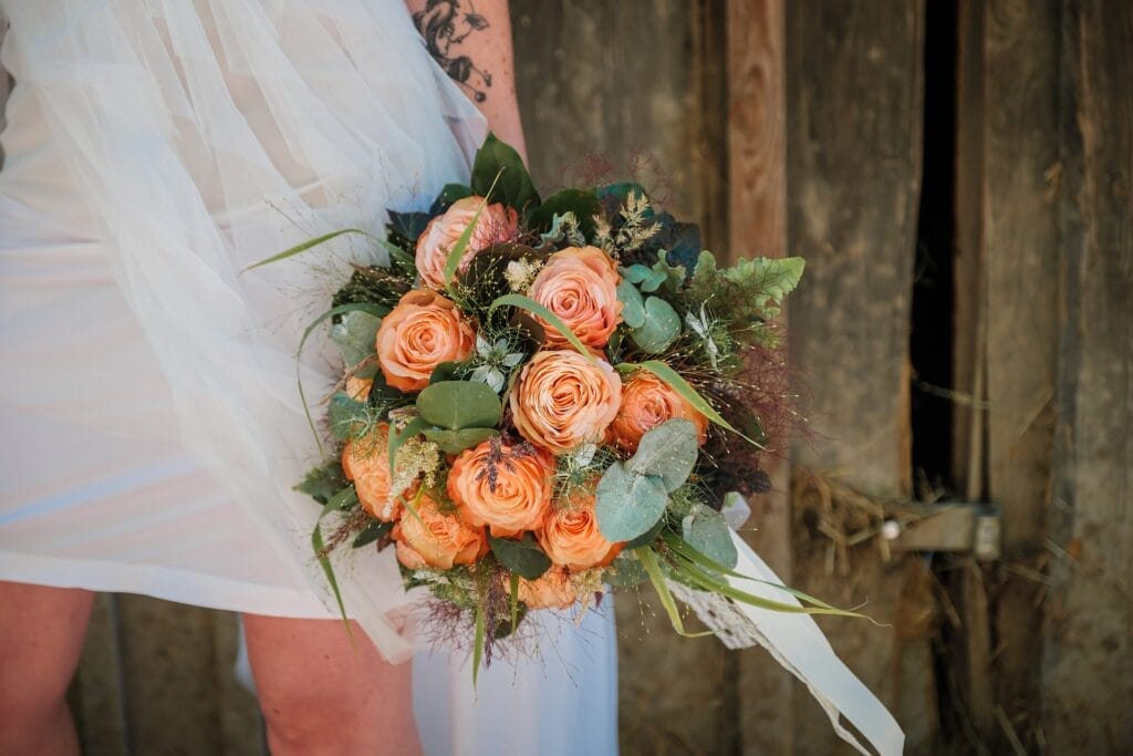 Hochzeitskrone Braut hält einen Strauß orangefarbener Rosen und Grünpflanzen vor einem rustikalen Holzhintergrund, eingefangen in echten Momenten von einem Hochzeitsfotografen in Wien und Niederösterreich.