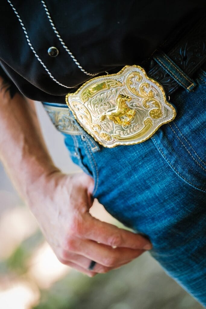 Hochzeitskrone Eine Person in Blue Jeans, schwarzem Hemd und einer großen, verzierten goldenen Gürtelschnalle mit Cowboy-Motiv fängt mühelos echte Momente ein.