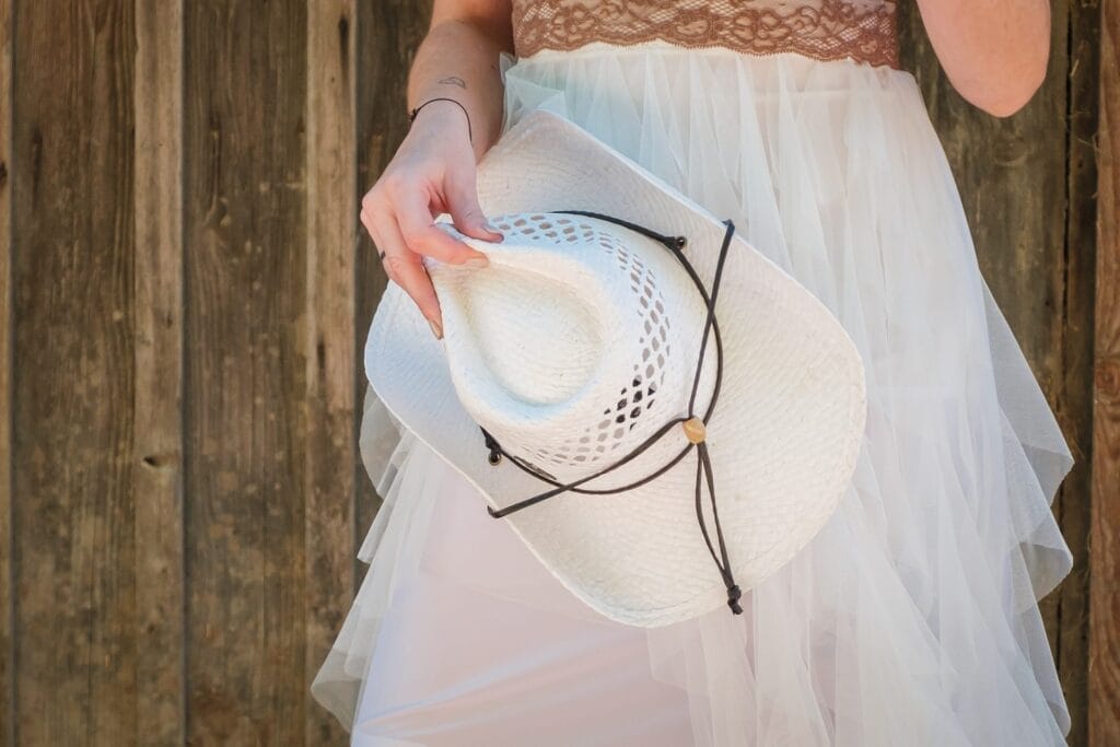 Hochzeitskrone Person in einer weißen Robe, die einen weißen Cowboy-Hut nahe einer Holz-Wand hält, eingefangen in echten Momenten. Diese natürlichen Hochzeitsfotos strahlen Authentizität aus und könnten von einem talentierten Hochzeitsfotografen in Wien und Niederösterreich stammen.