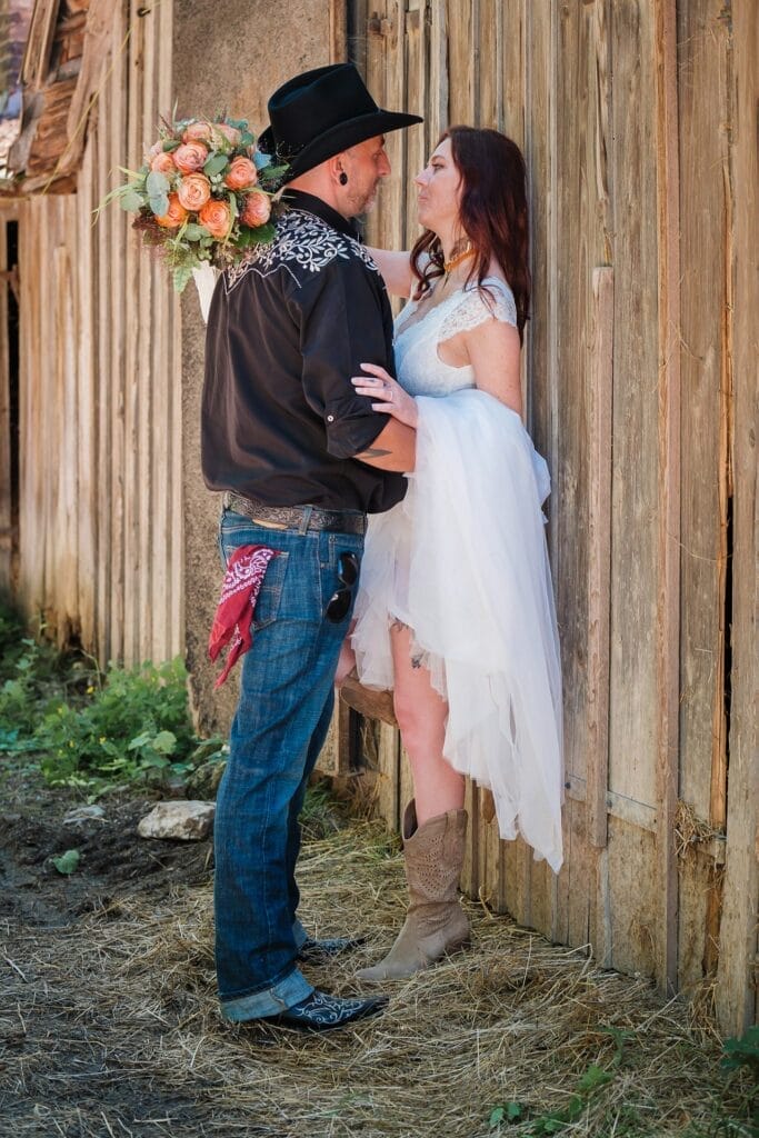 Hochzeitskrone Mann und Frau im Hochzeitskleid teilen einen intimen Moment an einer rustikalen Holzwand. Der Mann trägt einen schwarzen Cowboyhut und versteckt einen Blumenstrauß hinter seinem Rücken, während die Frau in weißen Stiefeln bezaubernd aussieht – echte Momente, wie sie nur natürliche Hochzeitsfotos einfangen können.