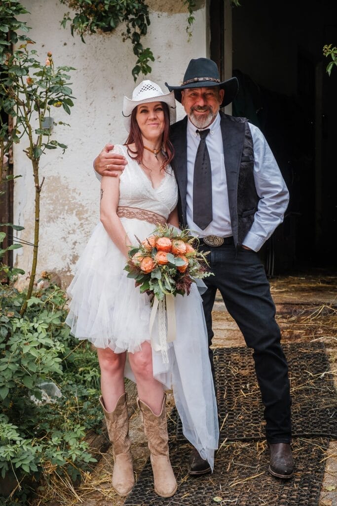 Hochzeitskrone Eine Frau in einem weißen Kleid und Cowboyhut hält einen Blumenstrauß und steht neben einem Mann mit Cowboyhut und Weste. Sie lächeln herzlich und fangen echte Momente der Freude vor einem rustikalen Gebäude ein.