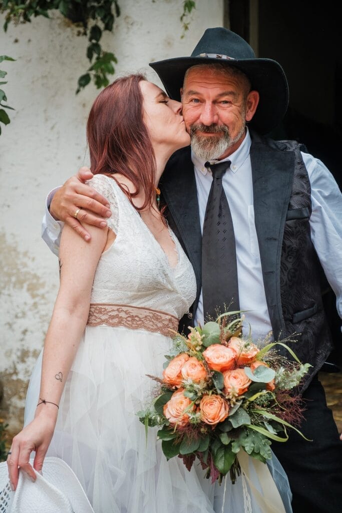 Hochzeitskrone Eine Frau im weißen Kleid küsst einen Mann mit Hut zärtlich auf die Wange; er trägt eine gemusterte Weste und Krawatte. Sie hält einen Strauß orangefarbener Rosen und Grünpflanzen in der Hand und fängt damit echte Momente ein. Perfekt für alle, die natürliche Hochzeitsfotos von einem Hochzeitsfotografen in Wien und Niederösterreich suchen.